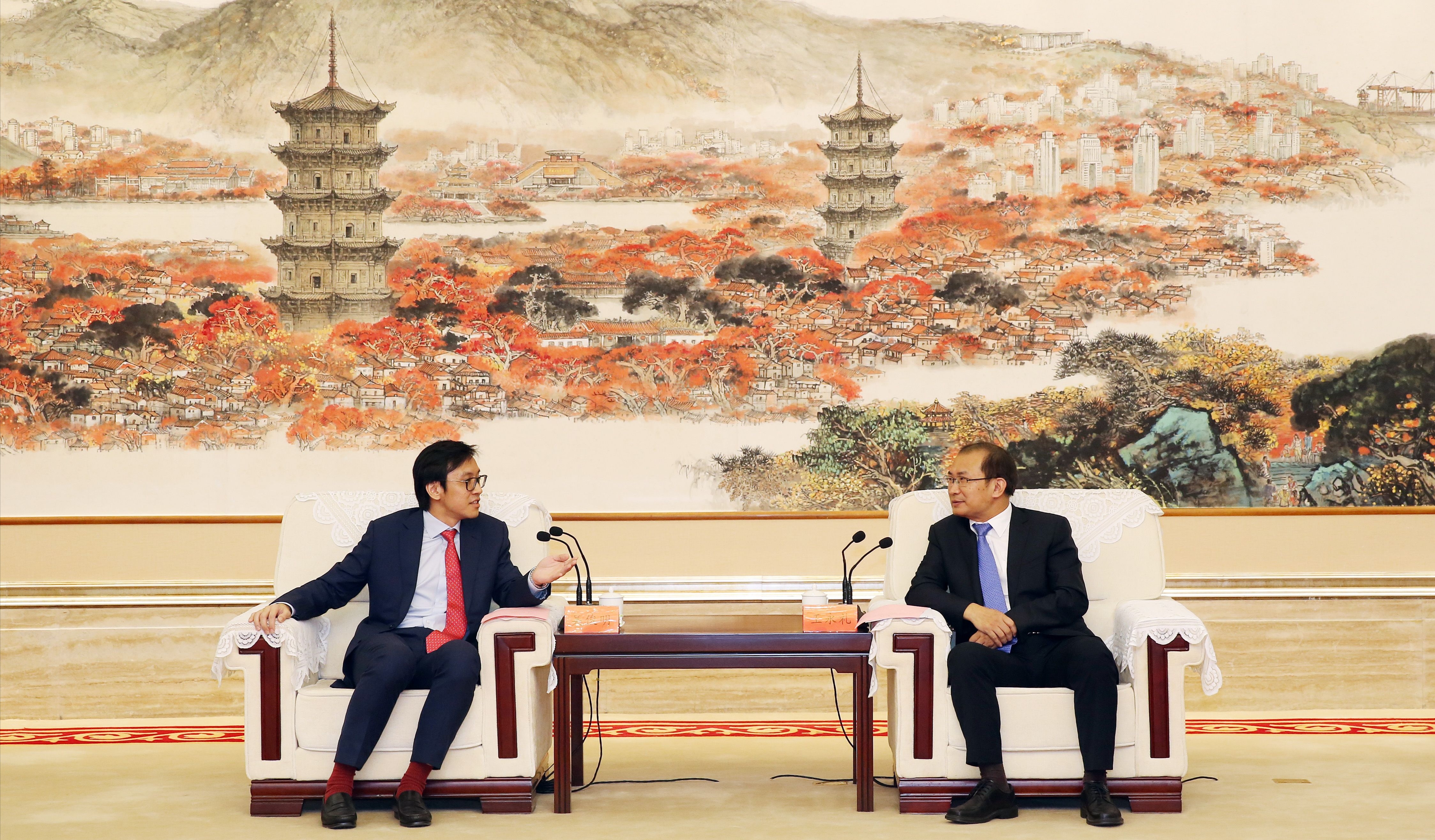 Two men in suits sit in armchairs facing each other, microphones between them, a landscape painting behind.